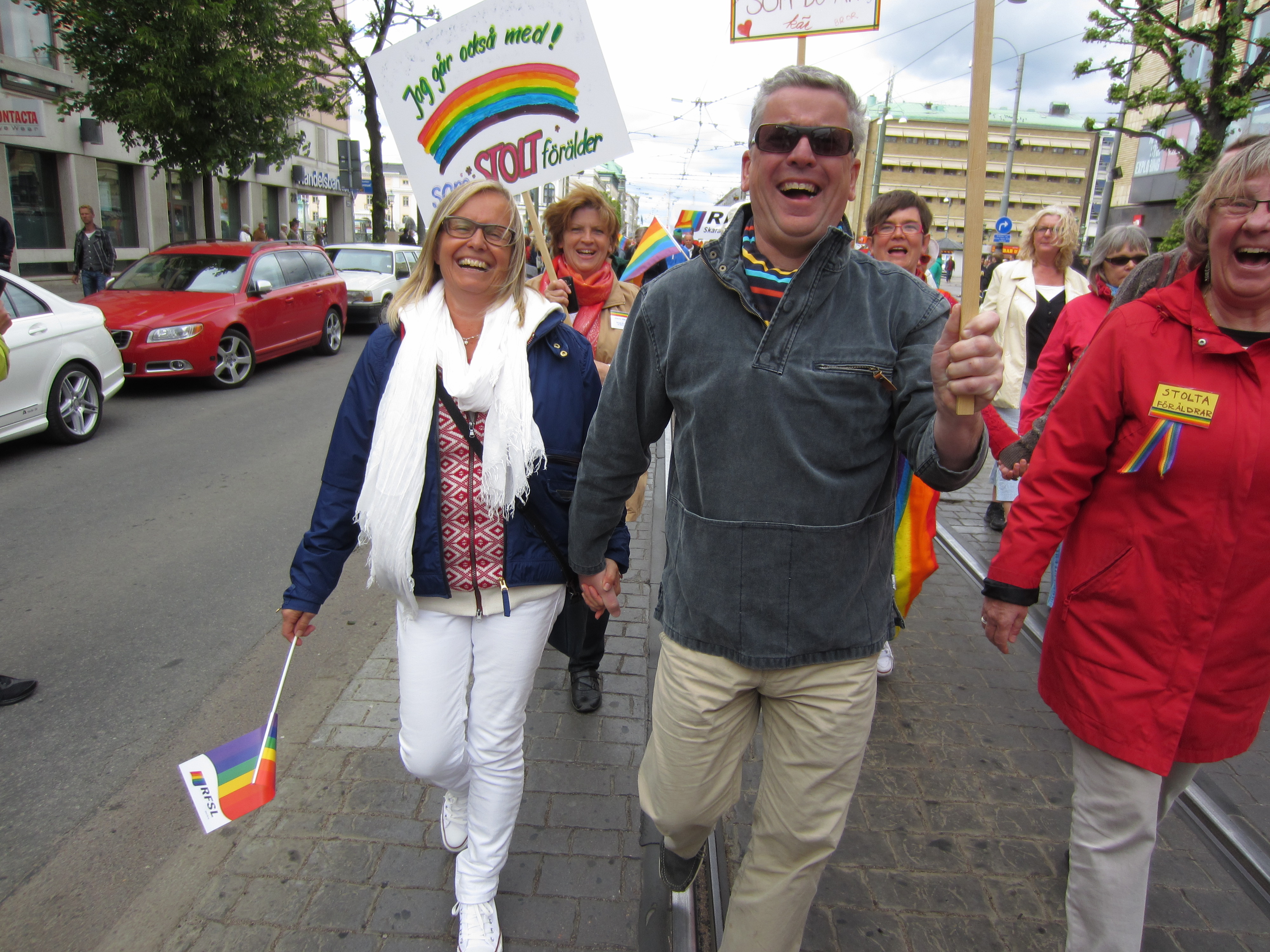 två stolta föräldrar håller varandra i händerna och ler stort mot kameran medan de går bland fler glada människor i pride parad med regnbågsflaggor och plakat